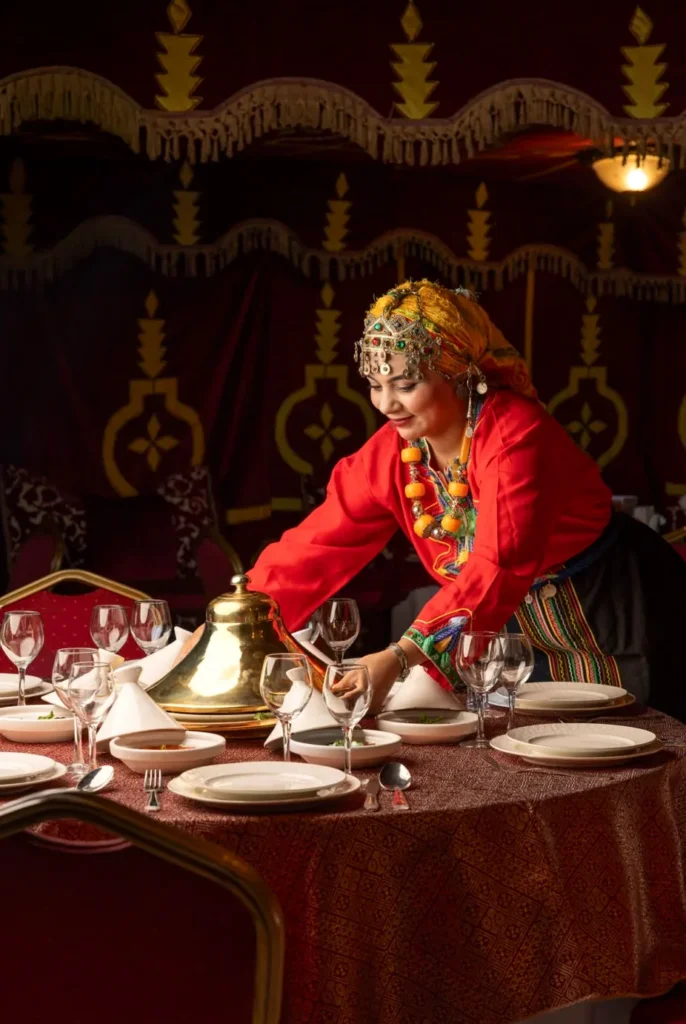 Woman in traditional Moroccan dress pouring tea at a dinner table under a red tent